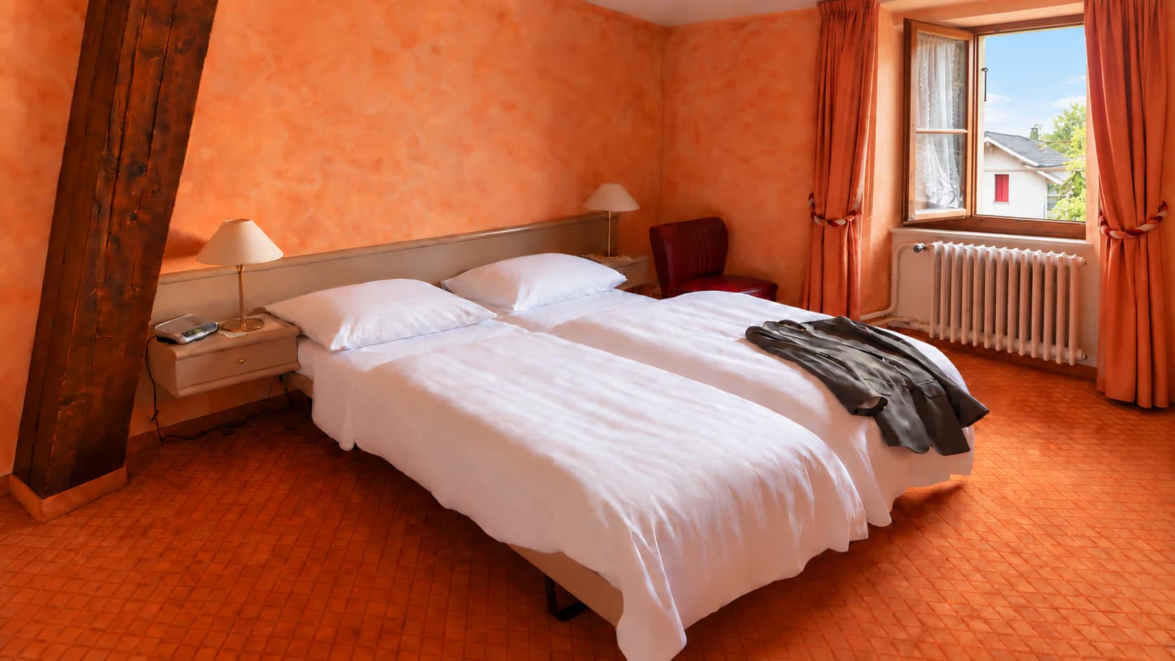 Chambre double aux murs orangés : lits jumeaux, fauteuil rouge et fenêtre ouverte sur le village